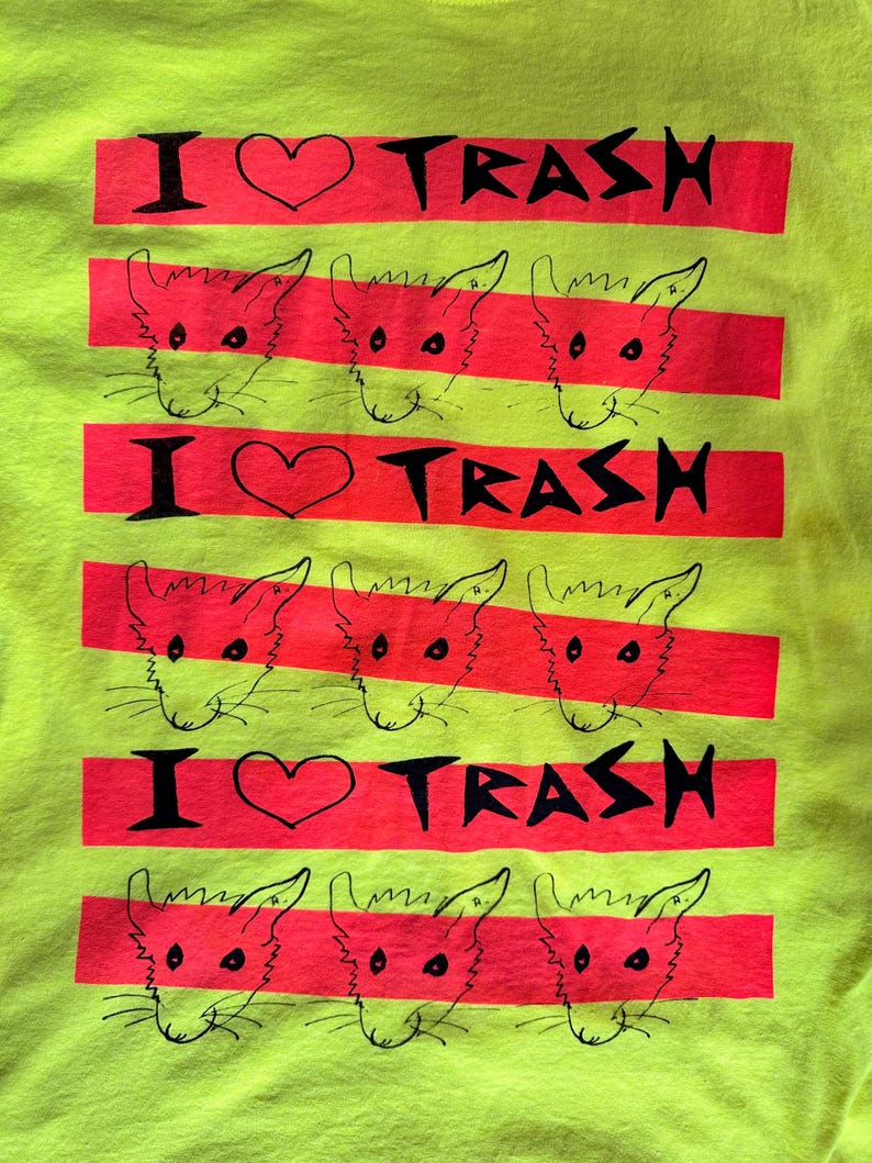 Pu&ograve; includere: T-shirt giallo neon con un design grafico. Il design presenta il testo "I ❤️ TRASH" in nero su una striscia orizzontale rossa, alternato a facce di gatti dei cartoni animati. La maglietta &egrave; audace e accattivante.