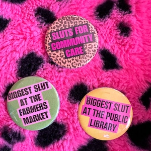 May include: Three round badges with text on a pink and black spotted background. One badge has a leopard print design with the text "SLUTS FOR COMMUNITY CARE". The other two badges say "BIGGEST SLUT AT THE FARMERS MARKET" and "BIGGEST SLUT AT THE PUBLIC LIBRARY".