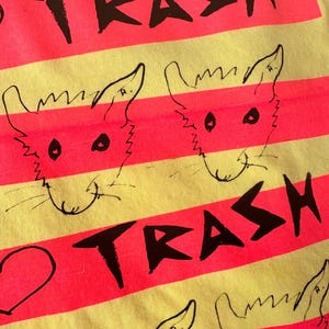 May include: Close-up of a yellow and pink striped t-shirt. The shirt features the word "TRASH" in black block letters and cartoon fox faces. The design is repeated across the fabric.