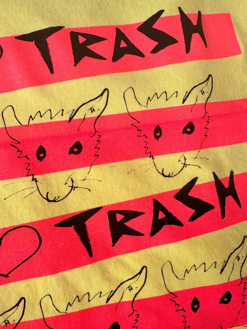 May include: Close-up of a yellow and pink striped t-shirt. The shirt features the word "TRASH" in black block letters and cartoon fox faces. The design is repeated across the fabric.