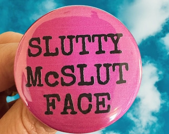 Può includere: Un bottone rosa con il testo "Slutty McSlut Face" in nero.