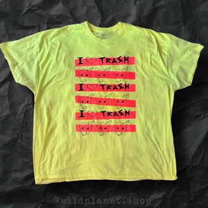 May include: A bright yellow t-shirt with a graphic design. The design features the text "I ❤️ TRASH" in black and red stripes, with rat illustrations. The shirt is made of a soft material.