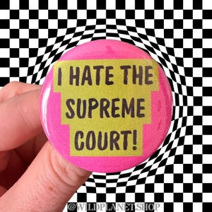 Peut inclure: Un bouton rose avec le texte "I HATE THE SUPREME COURT!" en noir et jaune. Le bouton est tenu devant un fond &agrave; carreaux noir et blanc. Le compte Instagram de la boutique, @WILDPLANETSHOP, est en bas.