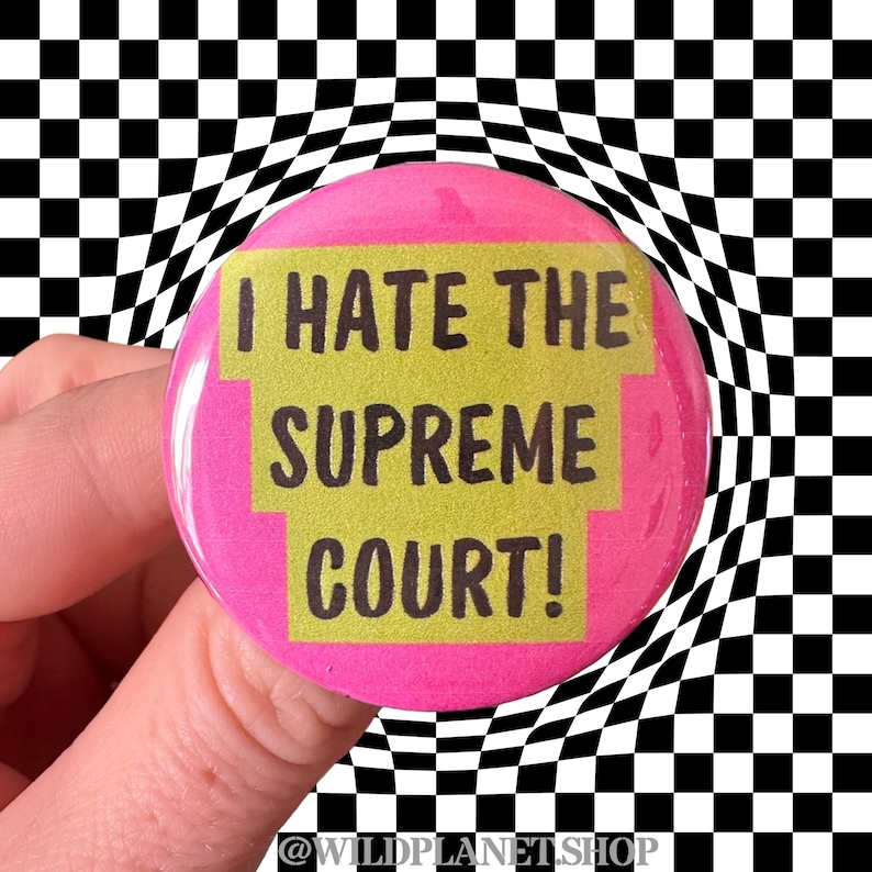 Peut inclure: Un bouton rose avec le texte "I HATE THE SUPREME COURT!" en noir et jaune. Le bouton est tenu devant un fond &agrave; carreaux noir et blanc. Le compte Instagram de la boutique, @WILDPLANETSHOP, est en bas.