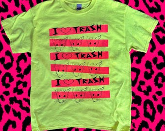 May include: A bright yellow t-shirt with a graphic design. The design features the text "I ❤️ TRASH" in black, with a heart symbol, and rat illustrations on red stripes. The shirt has a crew neck and short sleeves.