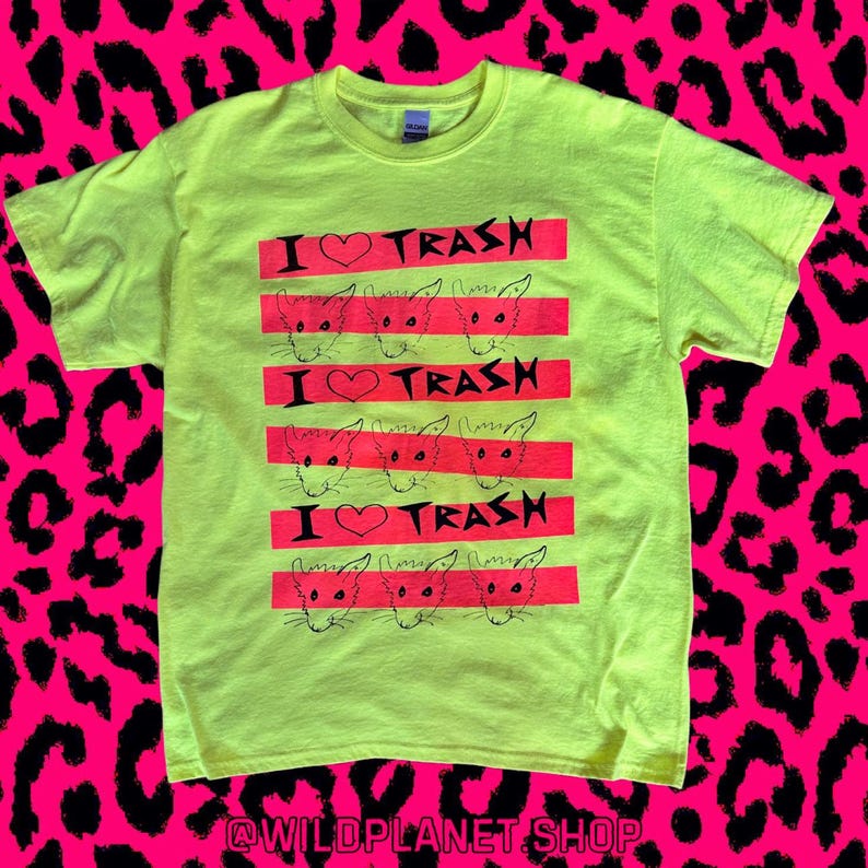 Pu&ograve; includere: Una maglietta giallo neon con un design grafico. Il design presenta il testo "I ❤️ TRASH" in nero, con un simbolo a cuore, e illustrazioni di ratti su strisce rosse orizzontali. La maglietta ha un girocollo e maniche corte.