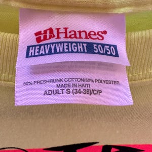 Pu&ograve; includere: Primo piano di un'etichetta di una maglietta giallo chiaro. L'etichetta &egrave; rosa con il logo Hanes rosso e le parole "HEAVYWEIGHT 50/50". Sotto il logo, l'etichetta recita "50% PRESHRUNK COTTON/50% POLYESTER MADE IN HAITI ADULT S (34-36)/C/P".