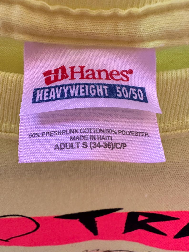 Pu&ograve; includere: Primo piano di un'etichetta di una maglietta giallo chiaro. L'etichetta &egrave; rosa con il logo Hanes rosso e le parole "HEAVYWEIGHT 50/50". Sotto il logo, l'etichetta recita "50% PRESHRUNK COTTON/50% POLYESTER MADE IN HAITI ADULT S (34-36)/C/P".