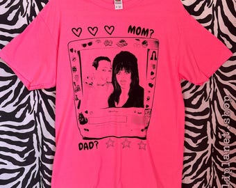 May include: A bright neon pink short-sleeve t-shirt featuring a black graphic print of a vintage television set displaying a man and a woman. The text MOM? appears above the screen and DAD? appears below, accompanied by heart and star icons.