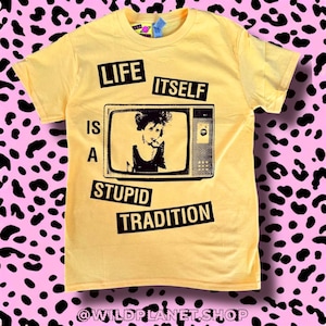 May include: Yellow t-shirt with the text "LIFE IS A STUPID TRADITION" in black block letters. A black and white image of a person on a vintage television screen is in the center. The shirt has short sleeves and a crew neck.