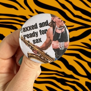 May include: A round button with a white background and a picture of a person playing a saxophone. The text on the button reads "axxed and ready for sax".