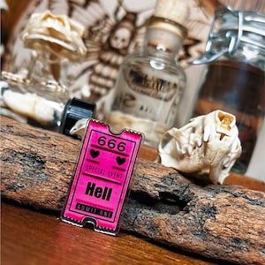 Könnte beinhalten: Ein rosa Ticket mit schwarzem Text, der "666 Special Event Hell Admit One" lautet. Das Ticket liegt auf einer Holzoberfläche mit einem Schädel und anderen Gegenständen.