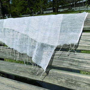May include: A white sheer scarf with a fringed edge draped over a wooden bench. The scarf is made of a lightweight, delicate fabric and has a subtle sheen.