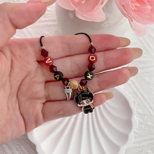 May include: A beaded bracelet featuring dark red faceted beads, black letter beads spelling VALOK, a single beige bead, a silver-toned tooth charm, and a black and white enamel charm depicting a stylized character with cat ears.