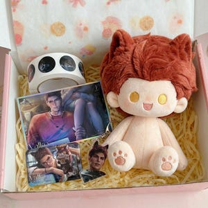 May include: A gift box containing a plush doll with rust-colored hair and cat ears, a roll of circular stickers, a rectangular card featuring a male character, and two small acrylic charms depicting a male character with cat ears.