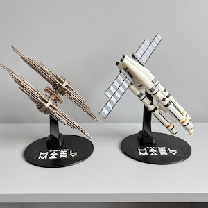 May include: Two detailed model spacecraft on black stands. One is metallic with multiple spires, the other is white with gray panels. Both have the text "PROJECT MARY" on their bases. The models are displayed on a white surface.