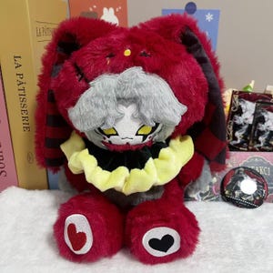 Può includere: Un peluche raffigurante un personaggio simile a un coniglio rosso con orecchie lunghe, capelli grigi e occhi gialli. Indossa un colletto arricciato giallo e nero. I piedi hanno toppe a forma di cuore, una rossa e una nera. Design cucito.