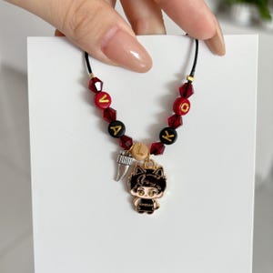 May include: A beaded bracelet on a black cord featuring red faceted crystal beads, black and red letter beads spelling VALKY, and a tan bead. The bracelet includes a silver-toned tooth charm and a gold-toned enamel charm of a cat-eared character.