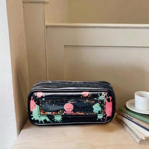 May include: A rectangular black pencil case with a clear front window containing loose colorful beads, a pink octopus figure, and a light blue squid figure. The exterior features black, pink, and light green paint splatter patterns.