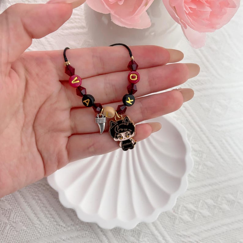 May include: A beaded bracelet featuring dark red faceted beads, black letter beads spelling VALOK, a single beige bead, a silver-toned tooth charm, and a black and white enamel charm depicting a stylized character with cat ears.