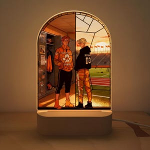 May include: An LED lamp with an illustration of two figures, one in a locker room, the other in a stadium at sunset. The lamp has a white base and emits a warm light. The design is framed by an arched shape.