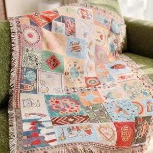 Könnte beinhalten: Eine gewebte Wohndecke mit einem Raster aus bunten Quadraten, die jeweils verschiedene Illustrationen wie Tiere, Himmelskörper, florale Muster und Symbole zeigen. Die Decke hat einen gefransten Rand und eine strukturierte, teppichartige Oberfläche.
