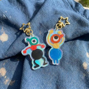 Puede incluir: Dos llaveros de acrílico con cierres dorados en forma de estrella. Uno presenta una figura abstracta verde, roja y negra, mientras que el otro muestra una figura naranja, amarilla y azul. Ambos dijes tienen un acabado iridiscente que refleja la luz.