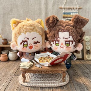 May include: Two plush dolls with anime-style faces, one with blonde hair and the other with brown hair, sit at a miniature table. A pizza box is open, revealing a pizza. A small stack of books and letters are also on the table.