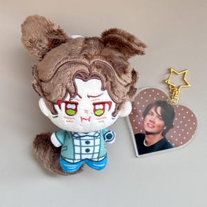 May include: A plush doll with brown fur and a cartoon face, wearing a teal jacket and blue pants. A heart-shaped keychain with a photo of a man is attached with a gold star-shaped clasp. The doll has a tail.