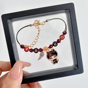 May include: A bracelet featuring a black cord, dark red faceted beads, and letter beads spelling VALKO. The piece includes a silver-toned fang charm and a small enamel charm depicting a stylized character with cat ears, secured by a gold-toned clasp.