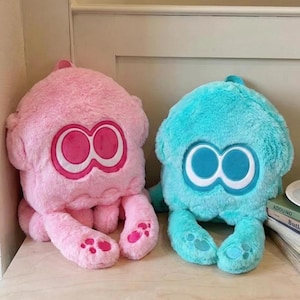 May include: Two plush backpacks shaped like squid characters, one in bright pink and one in light blue. Each features a soft, fuzzy texture, large embroidered eye patches with white outlines, and two dangling tentacles with embroidered paw-like details.