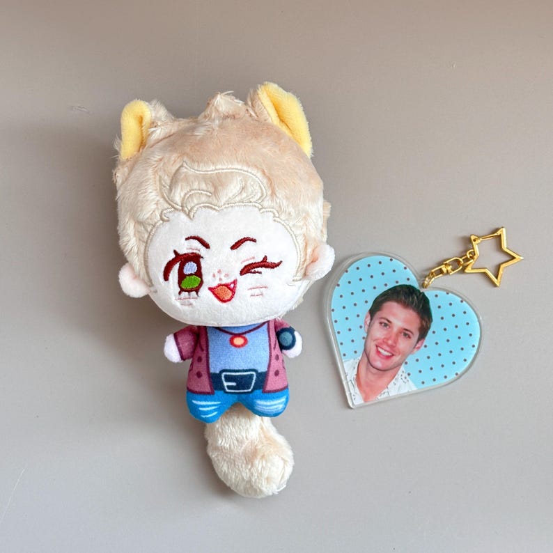 May include: A plush doll with a cartoon face, wearing a blue shirt, pink jacket, and blue pants. A heart-shaped keychain with a photo of a man is attached with a gold star charm. The doll has a light brown fur.