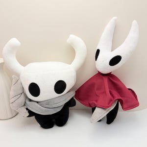May include: Two plush toys of video game characters. One is white with large horns, black eyes, a gray scarf, and a gray wing. The other is white with a red cape, black eyes, and a gray tail. Both have black bodies.