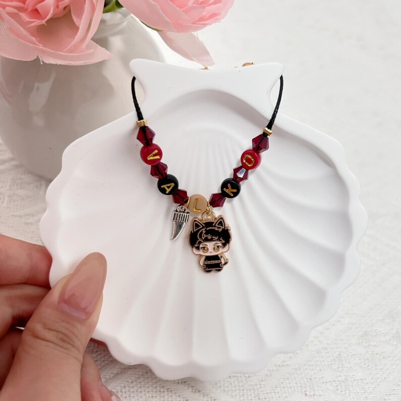 May include: A black cord bracelet featuring red and black beads with gold letters spelling VALKY. The bracelet includes a silver-toned tooth charm and a gold-toned enamel charm of a cartoon character with cat ears and black hair.