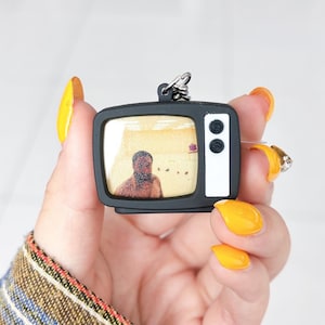Peut inclure: Un porte-clés miniature en forme de télévision noire avec un écran affichant une image granuleuse d'une personne à barbe foncée sur fond jaune. La télévision possède un panneau latéral blanc avec deux boutons noirs et un anneau en métal.