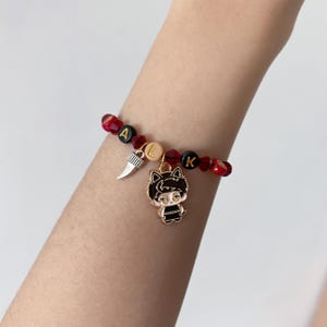 May include: A beaded bracelet featuring alternating red faceted beads and black round beads with gold lettering spelling WALK. The bracelet includes a silver-toned tooth charm and a gold-toned enamel charm depicting a stylized cat-eared character.