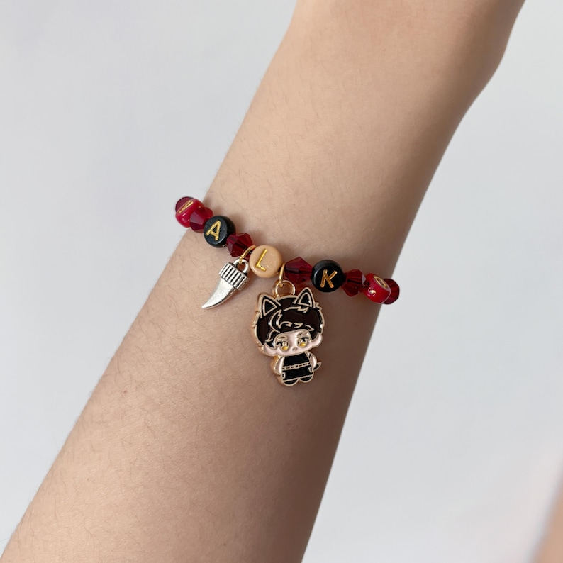 May include: A beaded bracelet featuring alternating red faceted beads and black round beads with gold lettering spelling WALK. The bracelet includes a silver-toned tooth charm and a gold-toned enamel charm depicting a stylized cat-eared character.