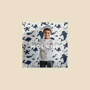 May include: A young person wearing a striped long-sleeve shirt and a gray beanie smiles in front of a wallpaper mockup. The wallpaper features a pattern of navy blue whales and fish on a white background. The words "WALLPAPER MOCKUP" are printed on the wallpaper.