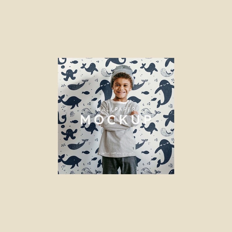 May include: A young person wearing a striped long-sleeve shirt and a gray beanie smiles in front of a wallpaper mockup. The wallpaper features a pattern of navy blue whales and fish on a white background. The words "WALLPAPER MOCKUP" are printed on the wallpaper.