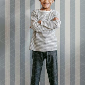 May include: A young person wearing a gray beanie, a striped long-sleeve shirt, and dark gray patterned pants. The person is smiling with arms crossed. The background features a striped wall.