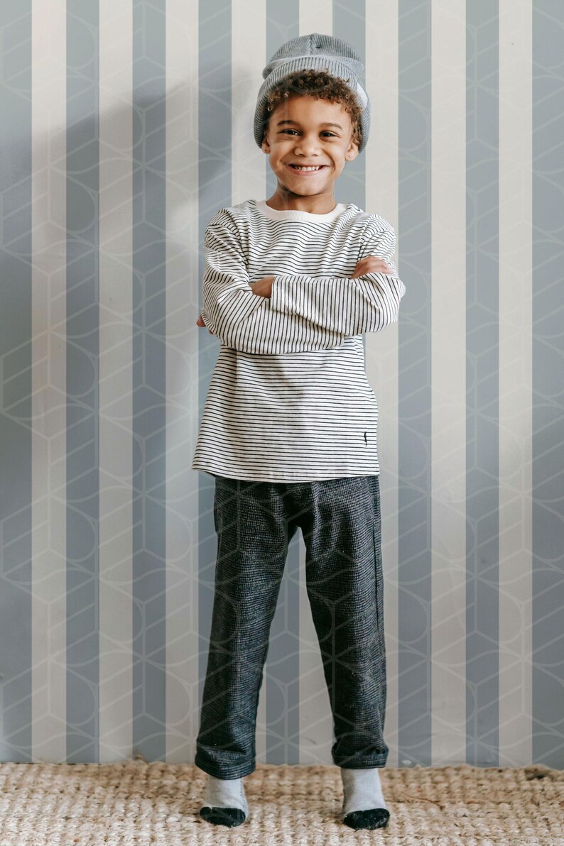 May include: A young person wearing a gray beanie, a striped long-sleeve shirt, and dark gray patterned pants. The person is smiling with arms crossed. The background features a striped wall.