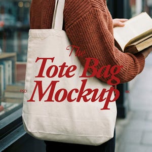 May include: A cream-colored tote bag with the words "The Tote Bag Mockup" in red script. The bag is held by a person wearing a rust-colored sweater. The bag has a simple design, with long straps for carrying.
