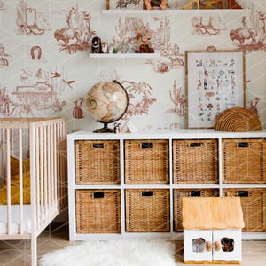 May include: A white crib with a yellow blanket and a white dresser with wicker baskets. The dresser is against a wall with a white and red wallpaper featuring a western theme. There is a globe, a framed alphabet chart, and a wooden toy house on the dresser.