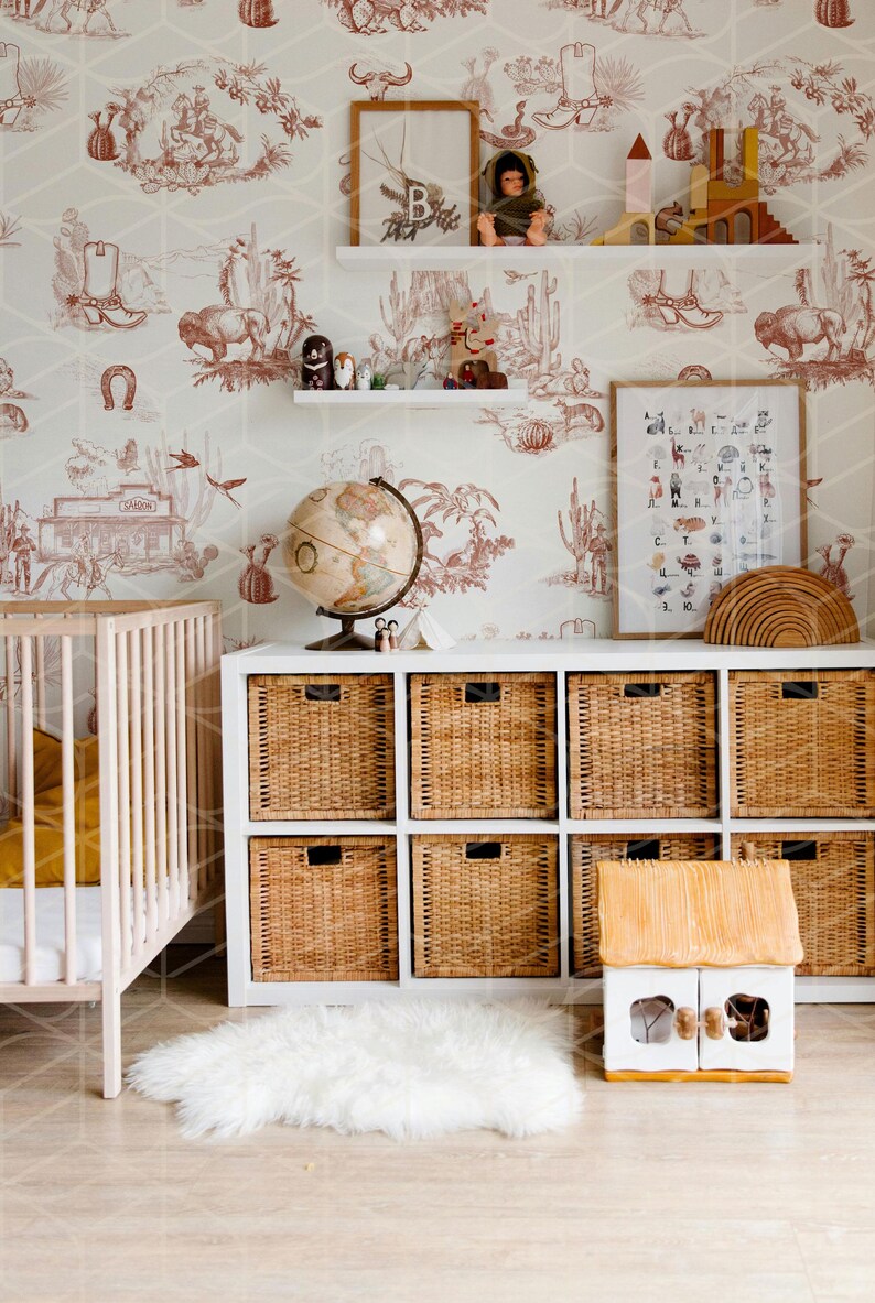 May include: A white crib with a yellow blanket and a white dresser with wicker baskets. The dresser is against a wall with a white and red wallpaper featuring a western theme. There is a globe, a framed alphabet chart, and a wooden toy house on the dresser.