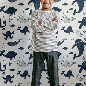 May include: A young person stands in front of a whimsical whale-themed wallpaper. They wear a striped long-sleeve shirt, patterned pants, and a gray beanie. The wallpaper features navy blue whales and fish on a light background.