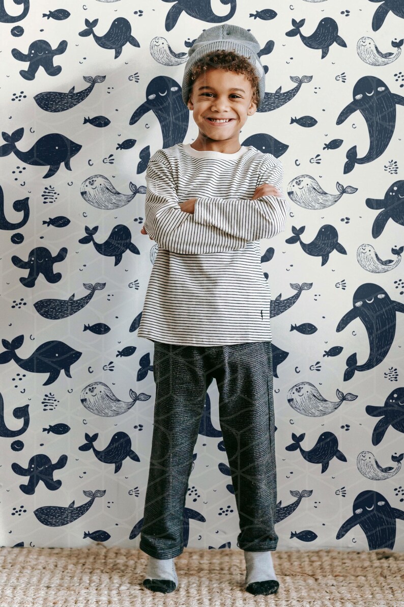 May include: A young person stands in front of a whimsical whale-themed wallpaper. They wear a striped long-sleeve shirt, patterned pants, and a gray beanie. The wallpaper features navy blue whales and fish on a light background.