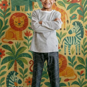May include: A child wearing a striped long-sleeve shirt, gray patterned pants, and a gray beanie smiles in front of a jungle-themed wallpaper. The wallpaper features illustrations of animals like lions, zebras, and elephants.