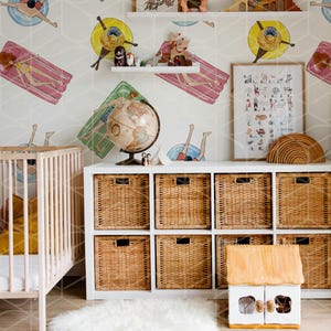 May include: A white crib with a yellow blanket and a white rug in front of a white cabinet with wicker baskets. The wall is covered in a wallpaper with a repeating pattern of people on inflatable rafts. There is a globe, a framed alphabet chart, and a wooden toy house on the shelves.