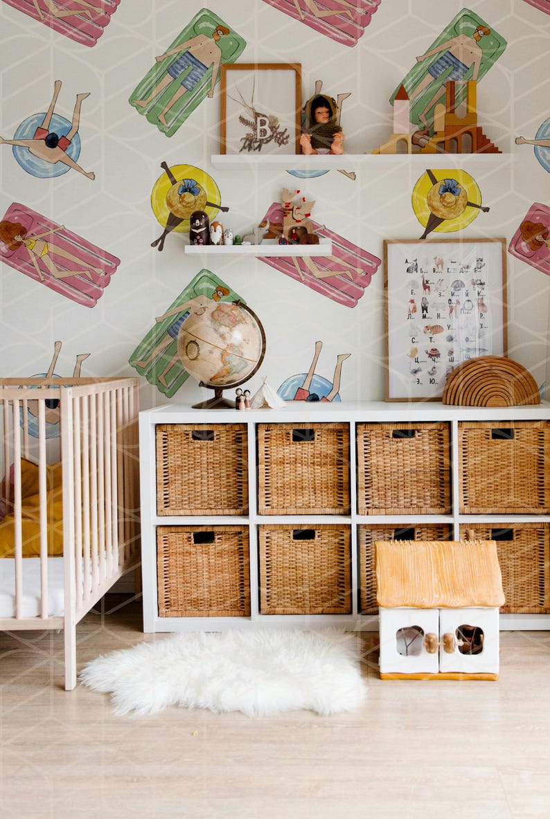 May include: A white crib with a yellow blanket and a white rug in front of a white cabinet with wicker baskets. The wall is covered in a wallpaper with a repeating pattern of people on inflatable rafts. There is a globe, a framed alphabet chart, and a wooden toy house on the shelves.
