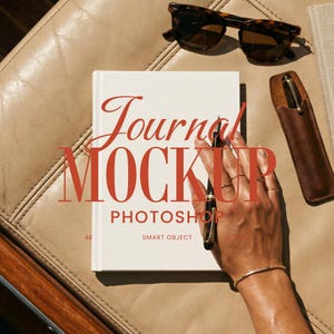 Puede incluir: Una maqueta de diario blanco con las palabras "Journal MOCKUP PHOTOSHOP SMART OBJECT" en rojo. La mano de una persona sostiene un bolígrafo sobre el diario. También se ven gafas de sol carey y un estuche de cuero.